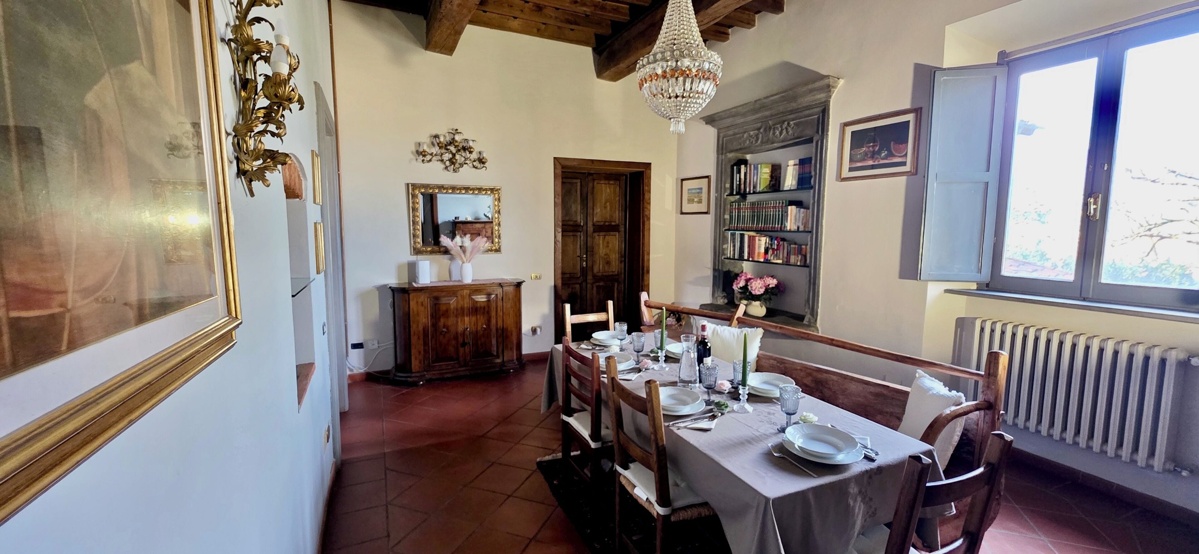 A dining room with a table set for four, a bookshelf, a mirror, and a chandelier.