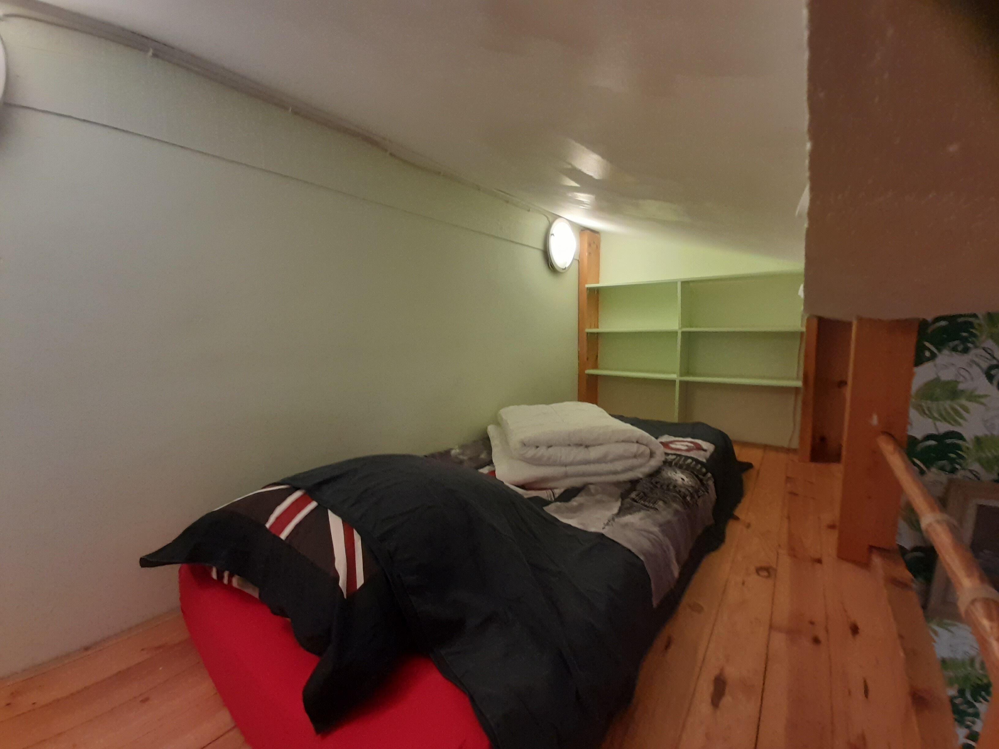 A small, narrow room with a single bed, a wooden floor, and a green shelf unit against the wall.