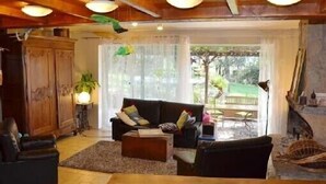 Een gezellige woonkamer met een houten plafond, een zwarte bank, een salontafel en uitzicht op de natuur door grote ramen.