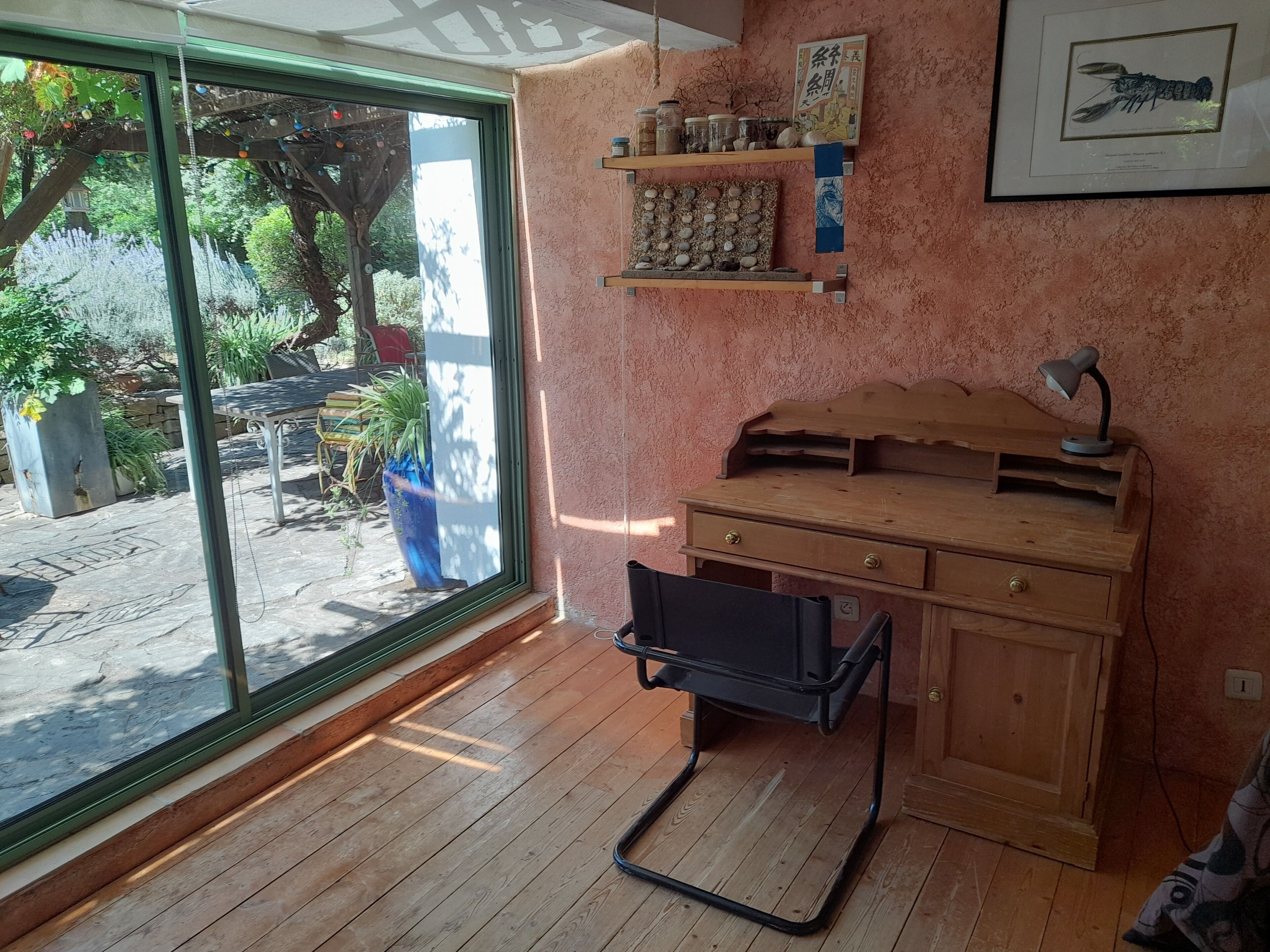 A room with a wooden desk, shelves with jars, a framed picture, and a large window overlooking a garden.