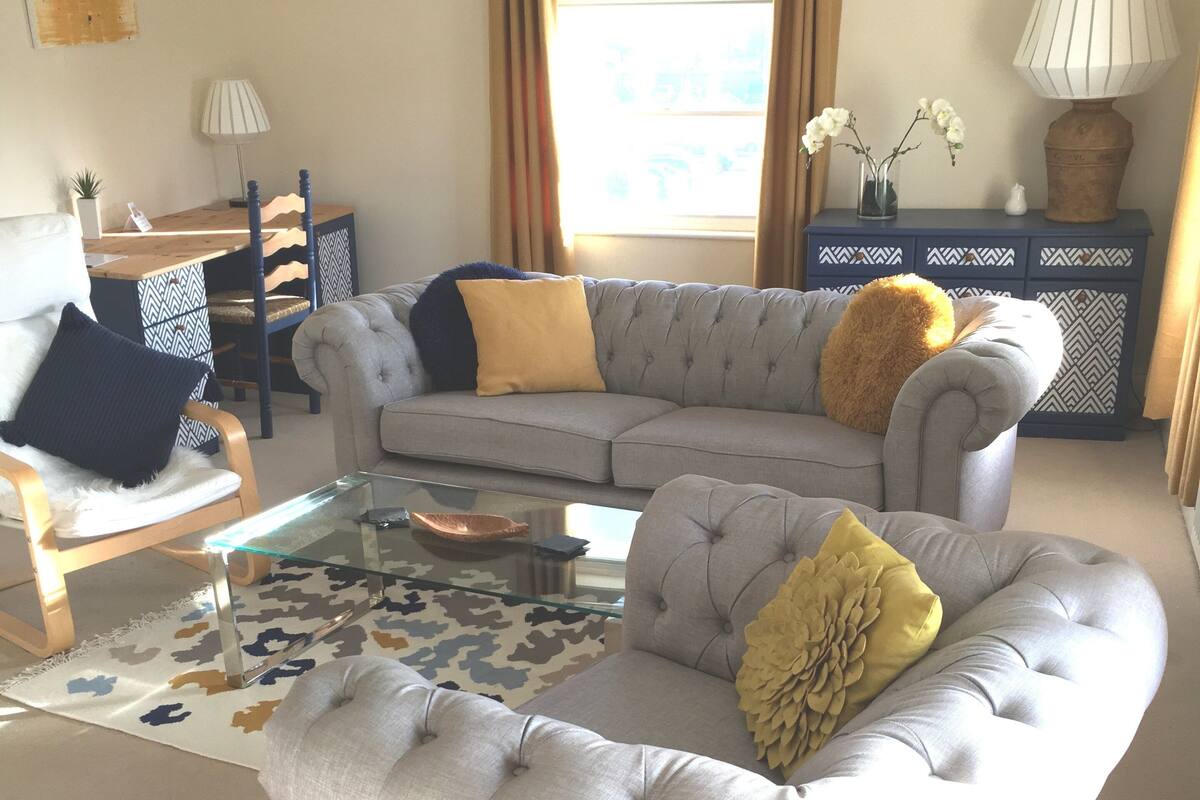 Un salon avec un canapé d’angle gris, une table basse en verre et un tapis à motifs.
