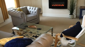 A living room with a beige sofa, a glass coffee table, a white armchair, and a fireplace. - Peel House Apartments (Burton on Trent)