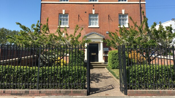 A red brick building with a central entrance, surrounded by a black metal fence and greenery. - Peel House Apartments (Burton on Trent)