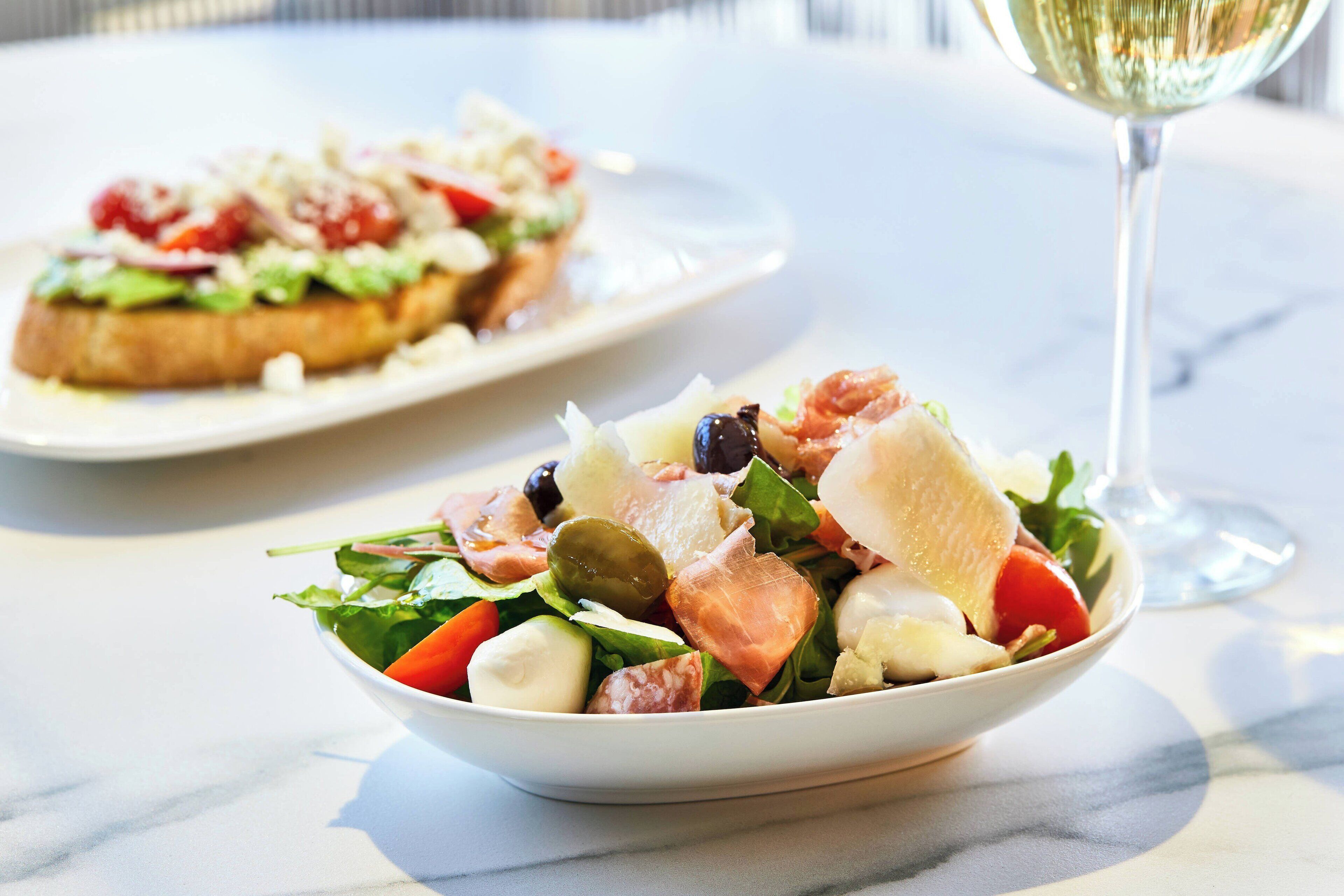 A bowl of salad with various ingredients and a plate of bruschetta in the background.