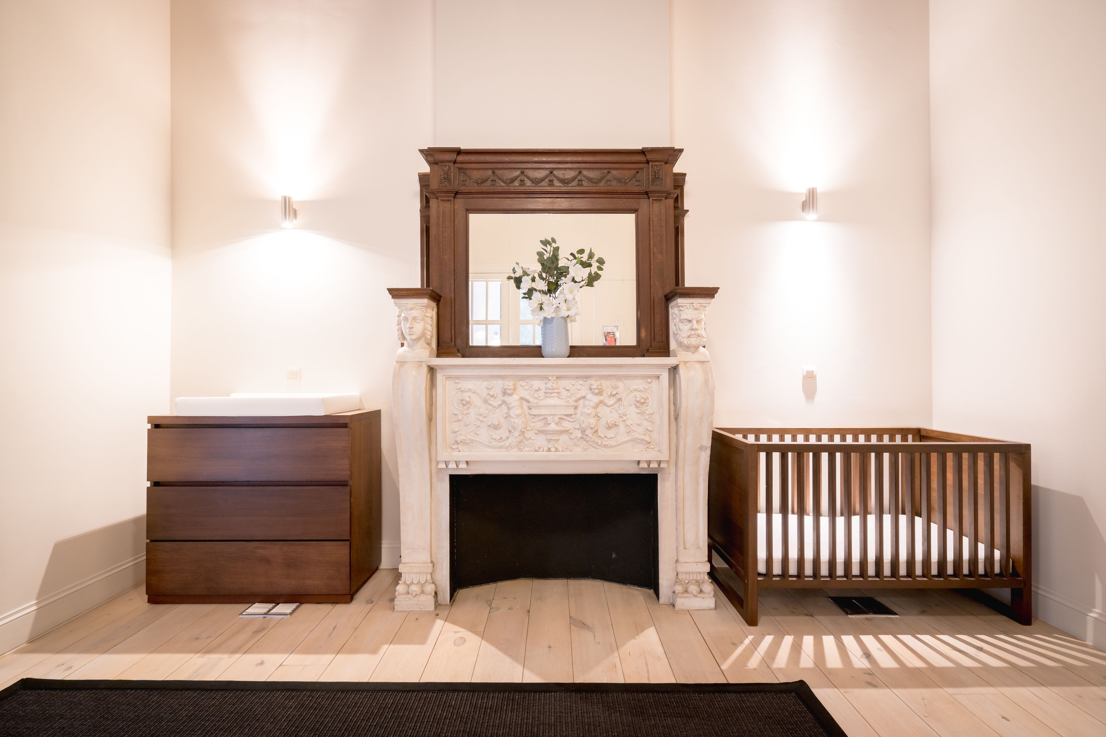 A baby room with a wooden crib, a wooden dresser, and a fireplace with a mirror above it.