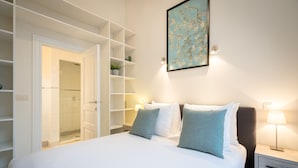 A hotel room with a bed, bedside tables, a nightstand, a wall-mounted lamp, a framed picture, and a built-in shelving unit. - Saint Pauls Suite 1 (Antwerp)