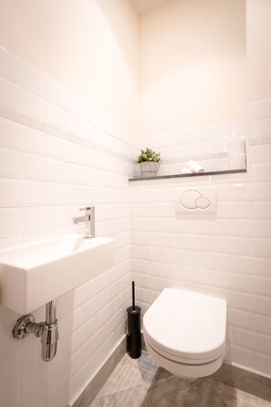 A bathroom with white tiled walls, a white sink, a toilet, and a small shelf with a plant and towels. - Saint Pauls Suite 1 (Antwerp)