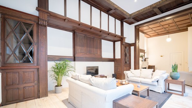 A spacious living room with a large white sofa, a wooden coffee table, and a television. There is a potted plant and a small table with decorative items. The room features exposed wooden beams and a large wooden door. - Saint Pauls Suite 1 (Antwerp)