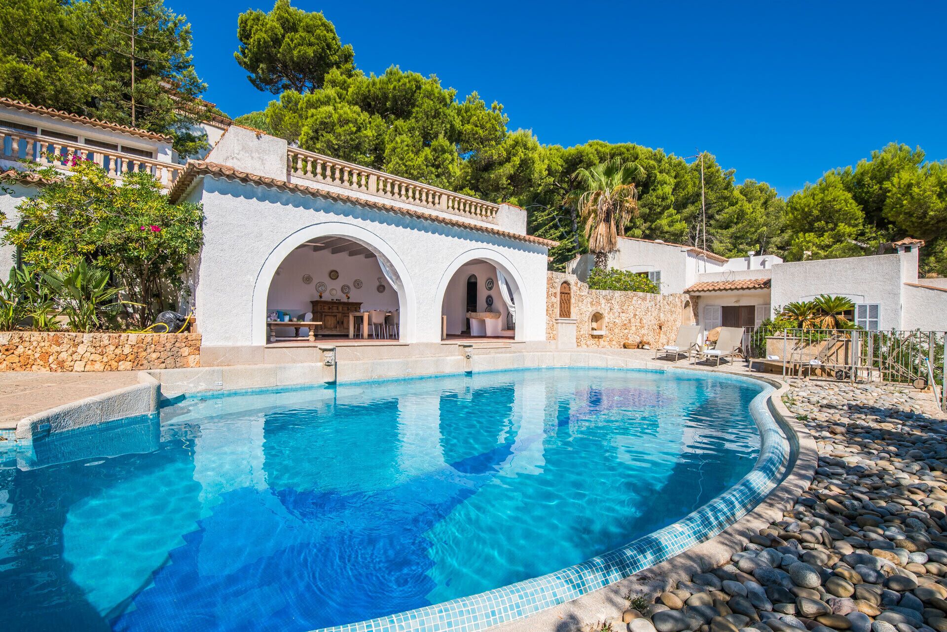 Una piscina con borde de piedra, un edificio blanco con arcos y vegetación circundante.