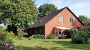 Une maison de plain-pied en briques, dotée d’un toit en pente, d’un patio couvert avec une table et des chaises, et d’un jardin bien entretenu où poussent diverses plantes et fleurs.