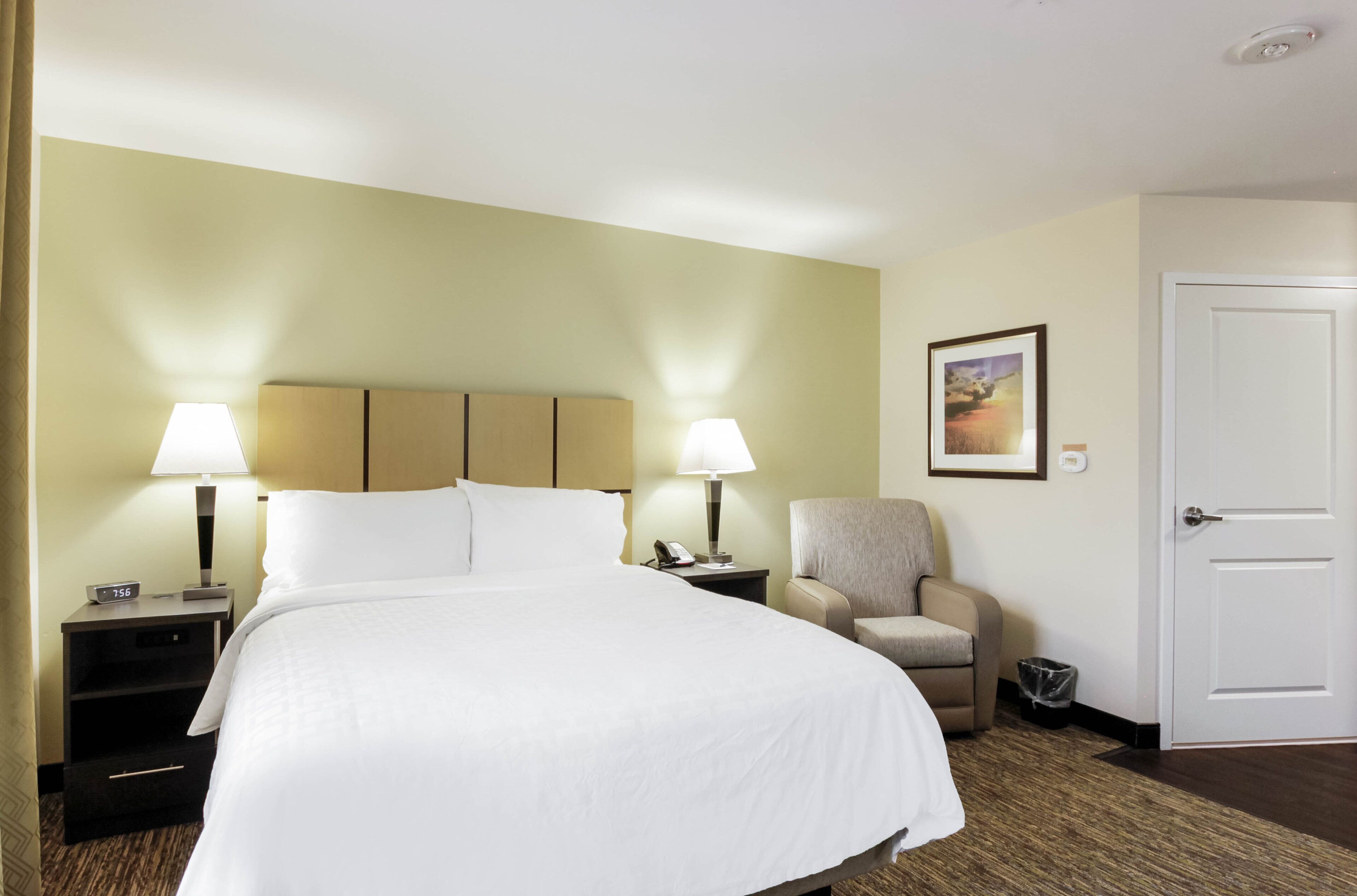 Una habitación de hotel con una cama grande, dos lámparas de noche, una silla, una mesita y una puerta.