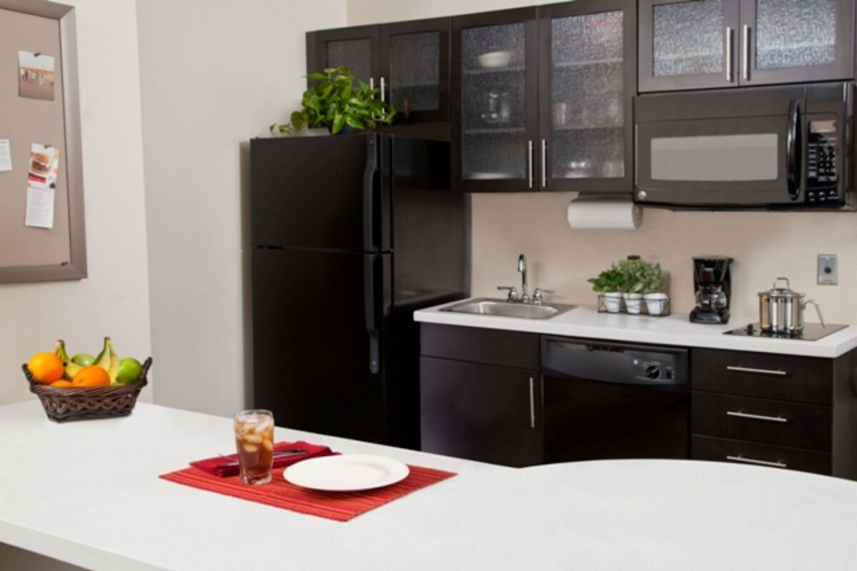 A modern kitchen with a black refrigerator, a microwave, and a sink. There is a fruit basket and a glass of iced tea on the counter.