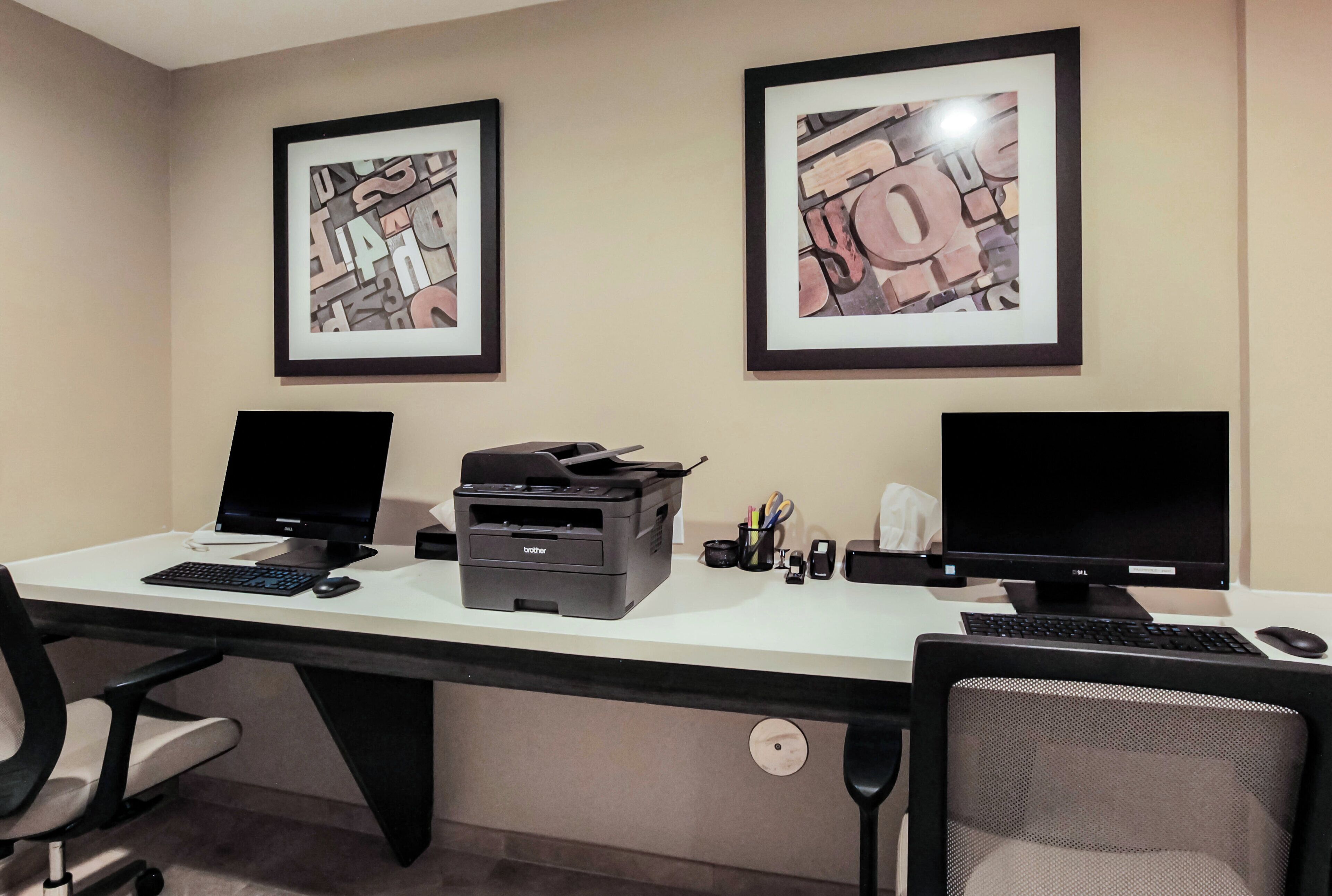 Een werkplek met twee schermen, een printer en kunstwerken in een lijst aan de muur.