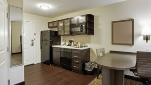 A compact kitchen with dark cabinets, a microwave, and a refrigerator. There's a small dining table with a chair. - Candlewood Suites Brighton by IHG (Brighton)