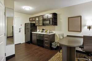 A compact kitchen with dark cabinets, a microwave, and a refrigerator. There's a small dining table with a chair. - Candlewood Suites Brighton by IHG (Brighton)