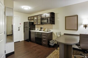 Cocina compacta con armarios oscuros, microondas y refrigerador. Hay una pequeña mesa de comedor con una silla.