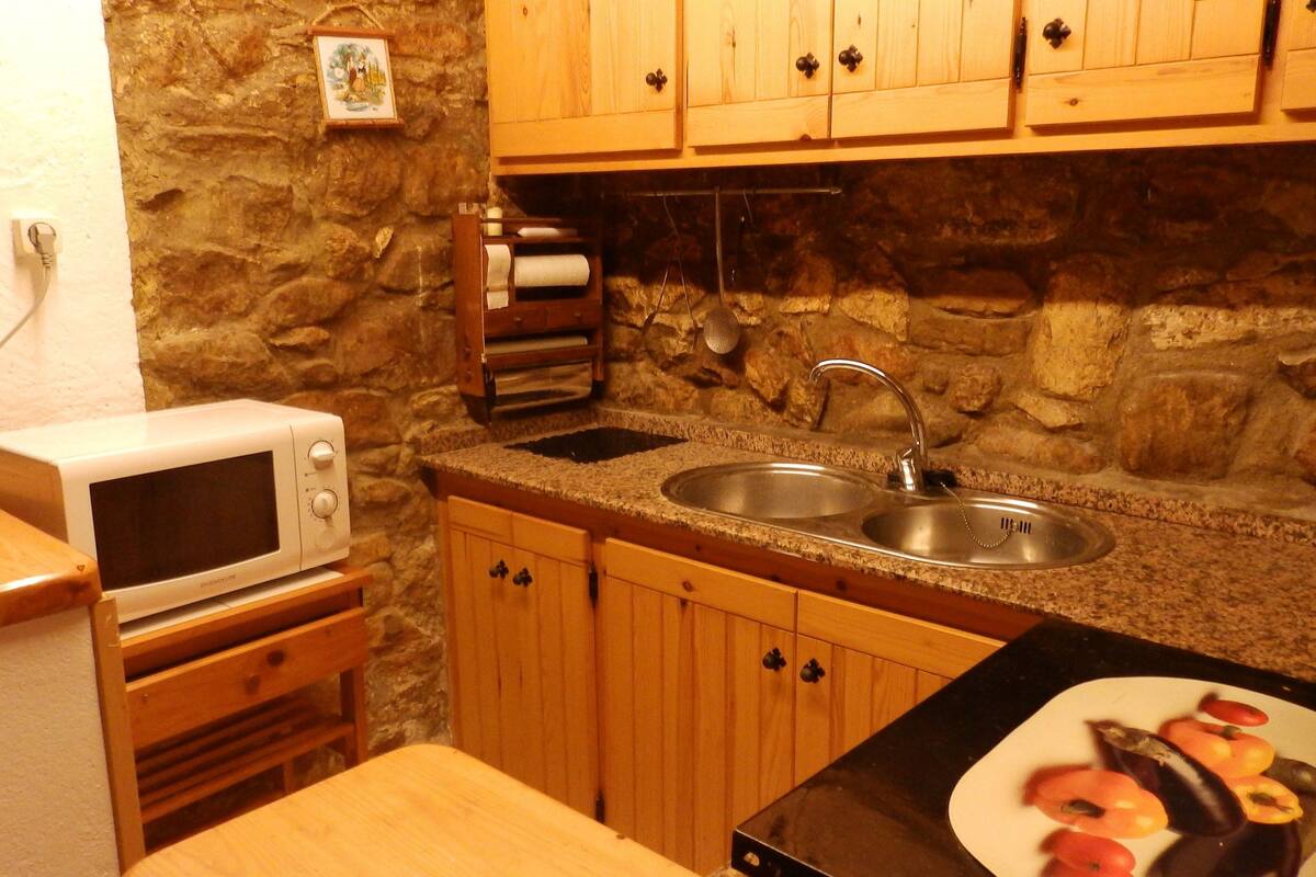 A compact kitchen with a stone wall, wooden cabinets, a microwave, a sink, and a cutting board with vegetables.