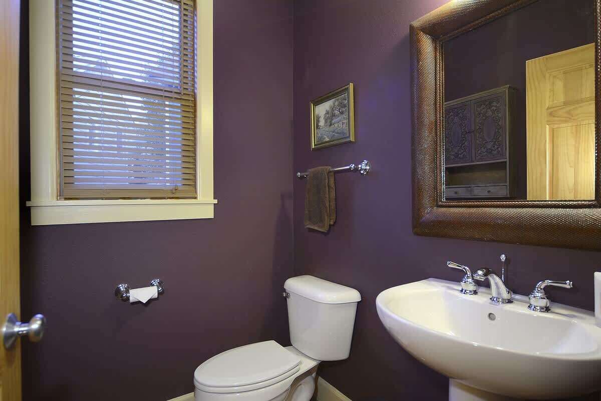 Une salle de bain comprenant des toilettes, un lavabo, une fenêtre avec stores, un porte-serviettes et un miroir avec un meuble de rangement au-dessus.