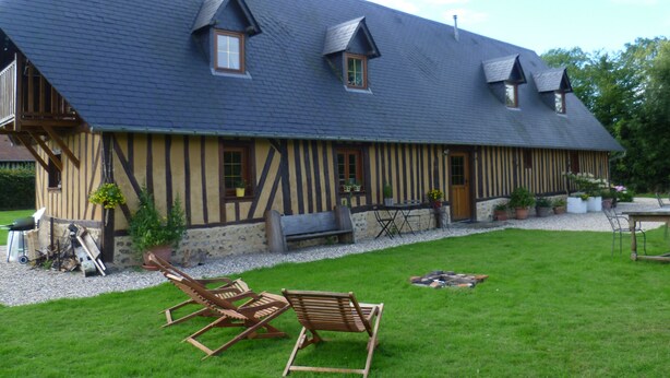 A traditional half-timbered house with a sloped roof, a small garden with lawn chairs, and a gravel pathway leading to the entrance.