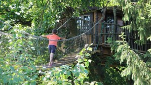 A person crossing a rope bridge in a forested area with a wooden structure in the background.