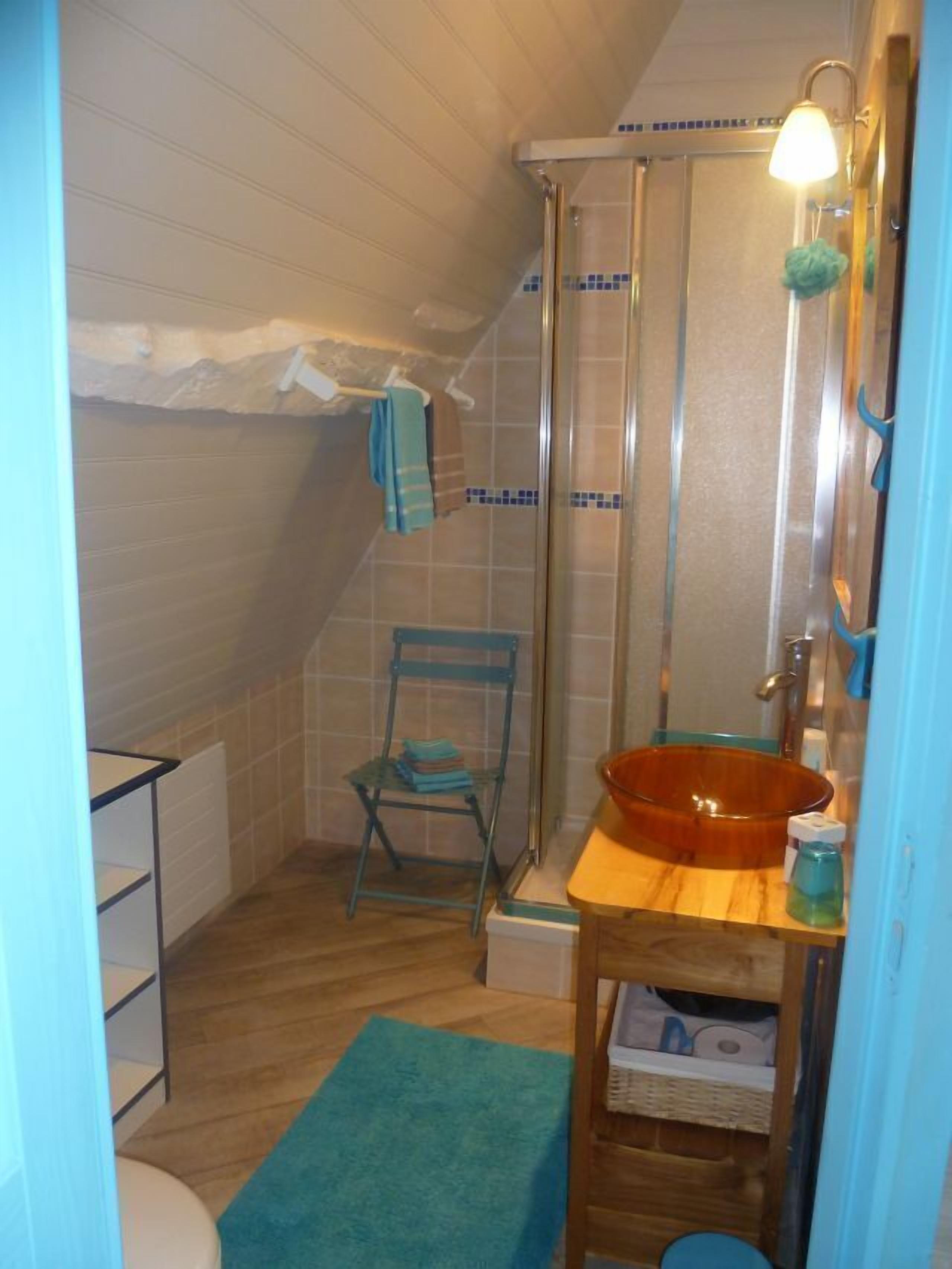 A small, compact bathroom with a shower, a sink with a bowl basin, a chair, and a shelf with toiletries.