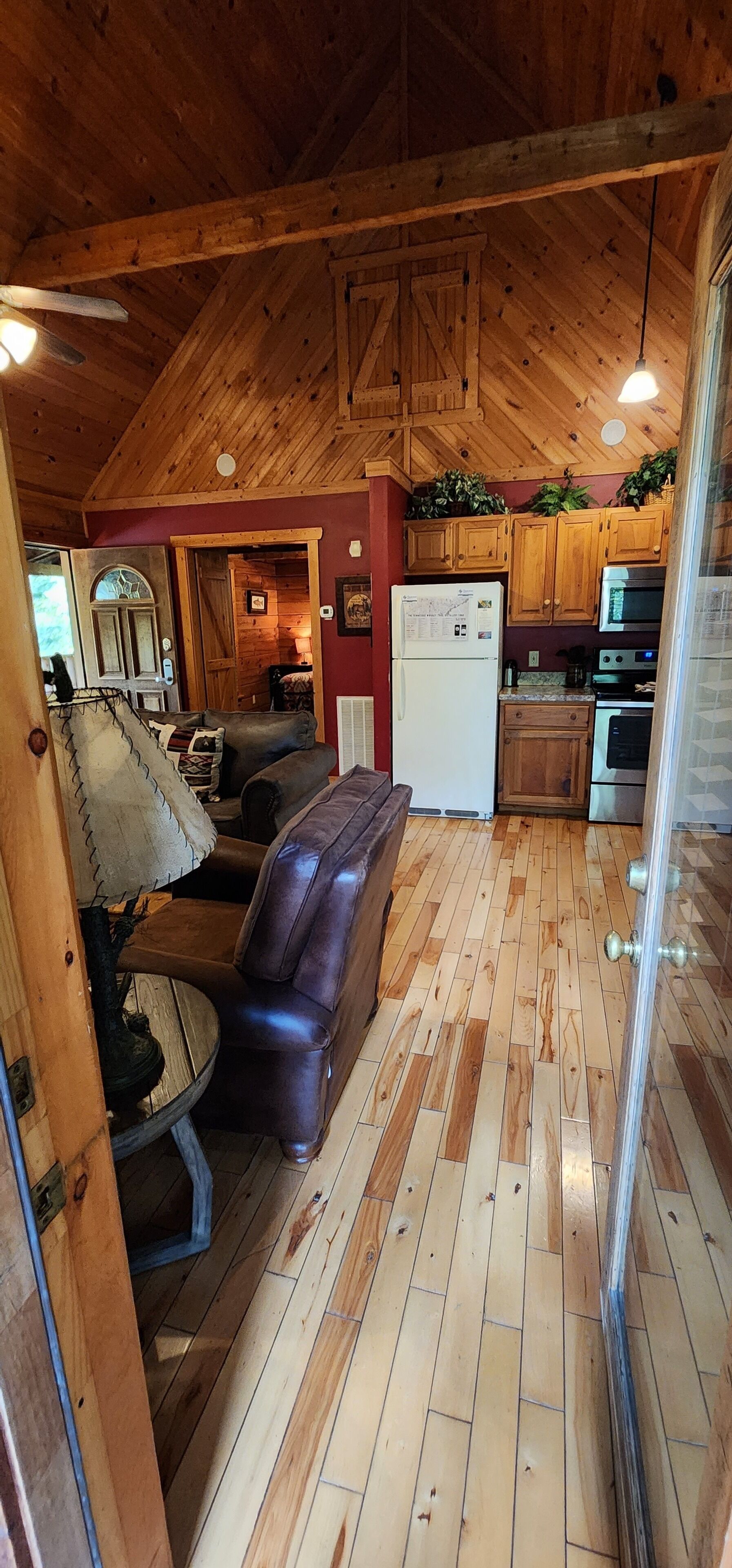 Un acogedor interior de cabaña con techo de madera, una sala de estar con sofá y lámpara, una cocina con armarios y electrodomésticos de madera, y un comedor con mesa y sillas.