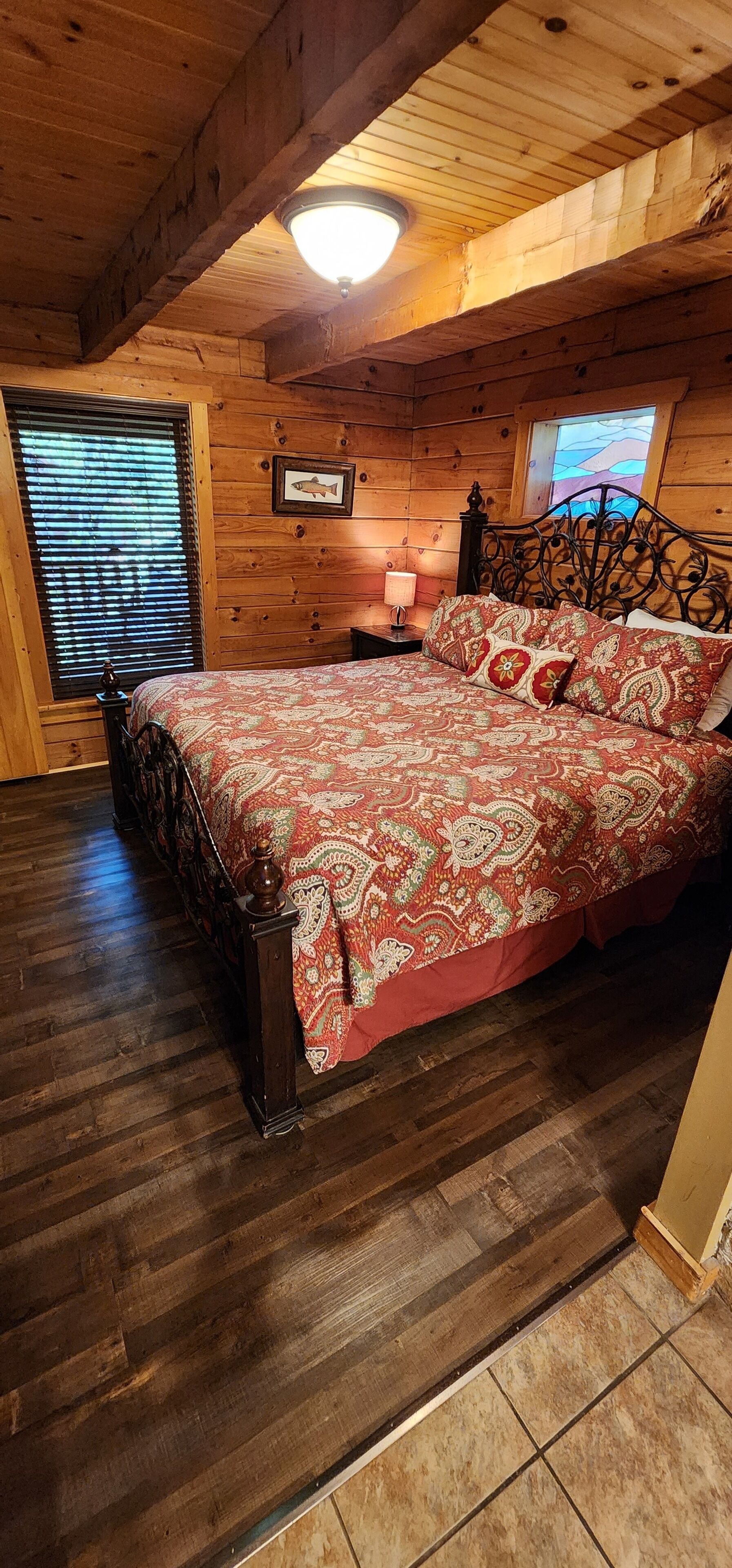 A wooden cabin bedroom with a bed, a nightstand, a lamp, and a window with blinds.