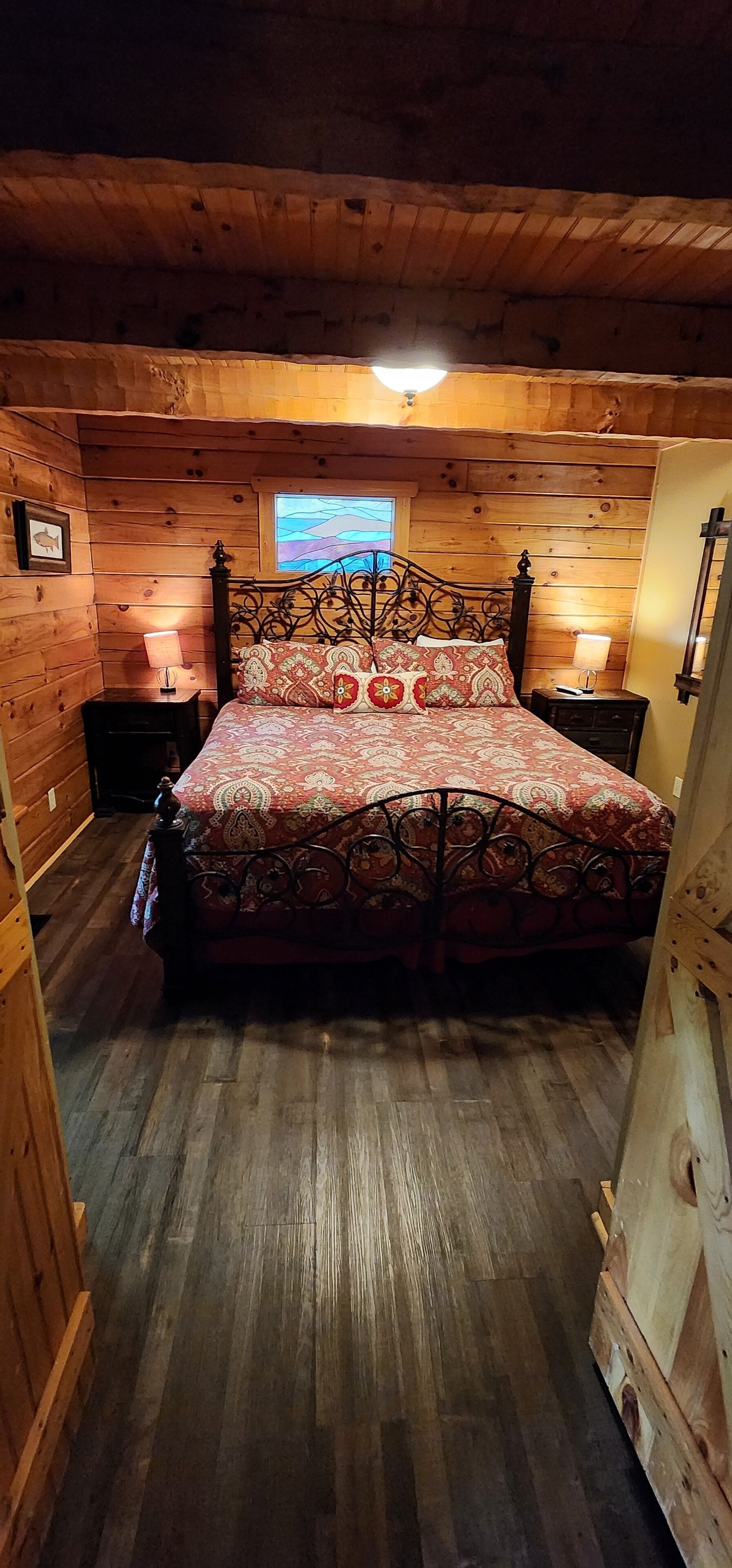 A wooden cabin bedroom with a bed, two nightstands, a window with a view, and a wooden floor.