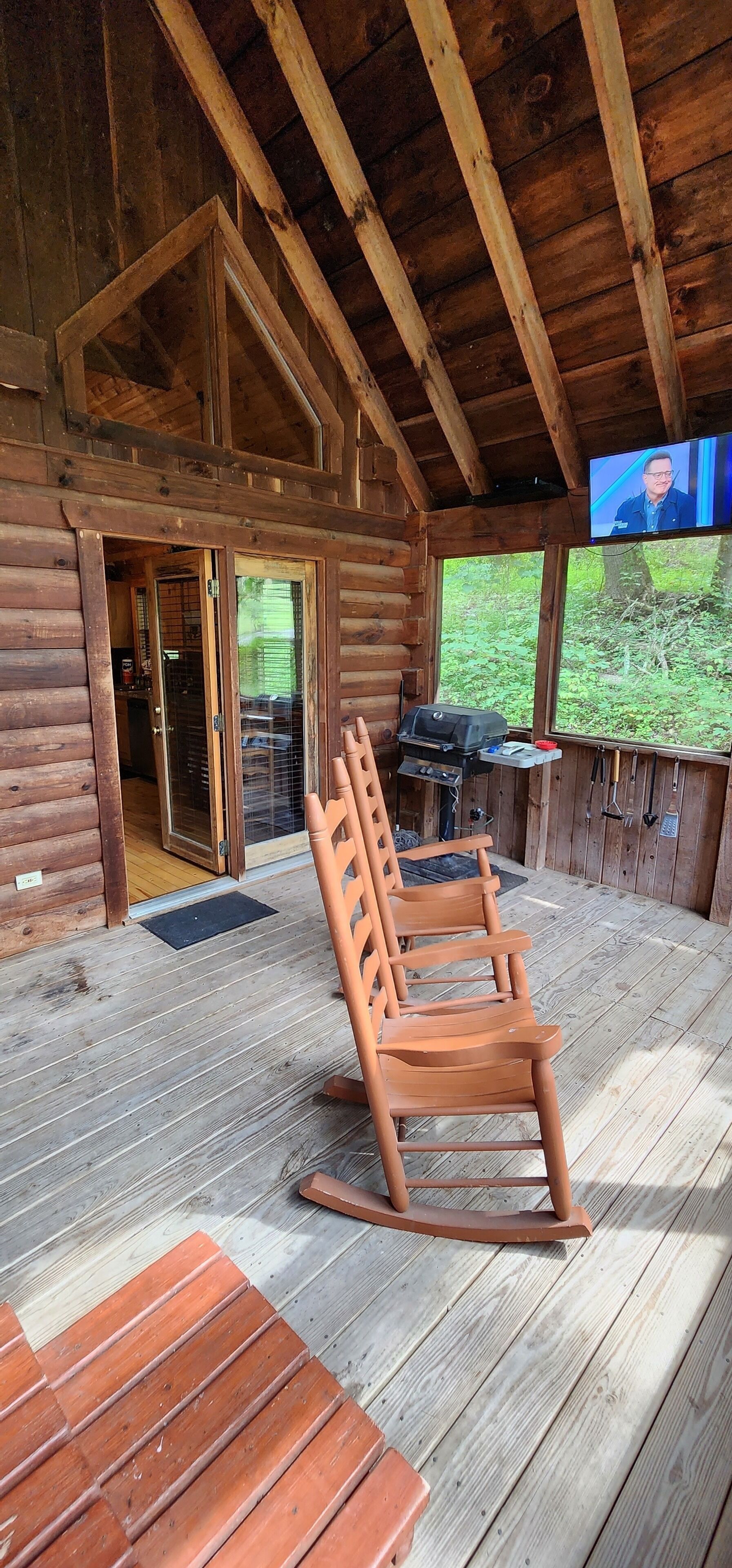 Uma cabana de madeira com varanda, porta de vidro deslizante, churrasqueira e televisão fixada na parede.
