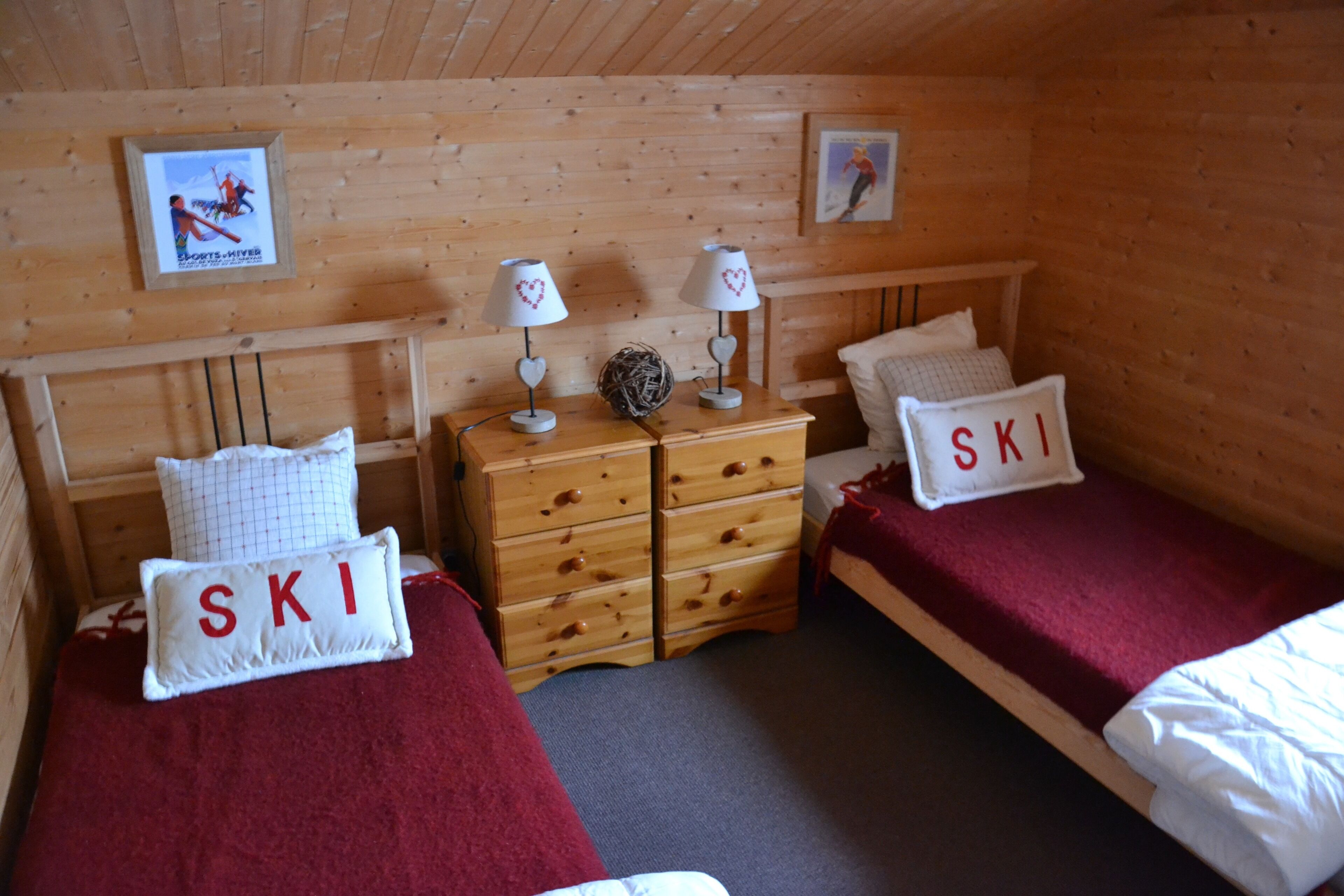 A wooden cabin room with two beds, each with a pillow labeled "SKI". There are two wall-mounted lamps with heart-shaped lampshades, a wooden dresser with drawers, and a framed picture on the wall.