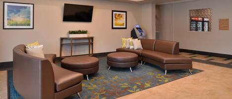 A lobby with a brown leather sofa, a round ottoman, a small table with a plant, a mounted TV, and a reception desk.