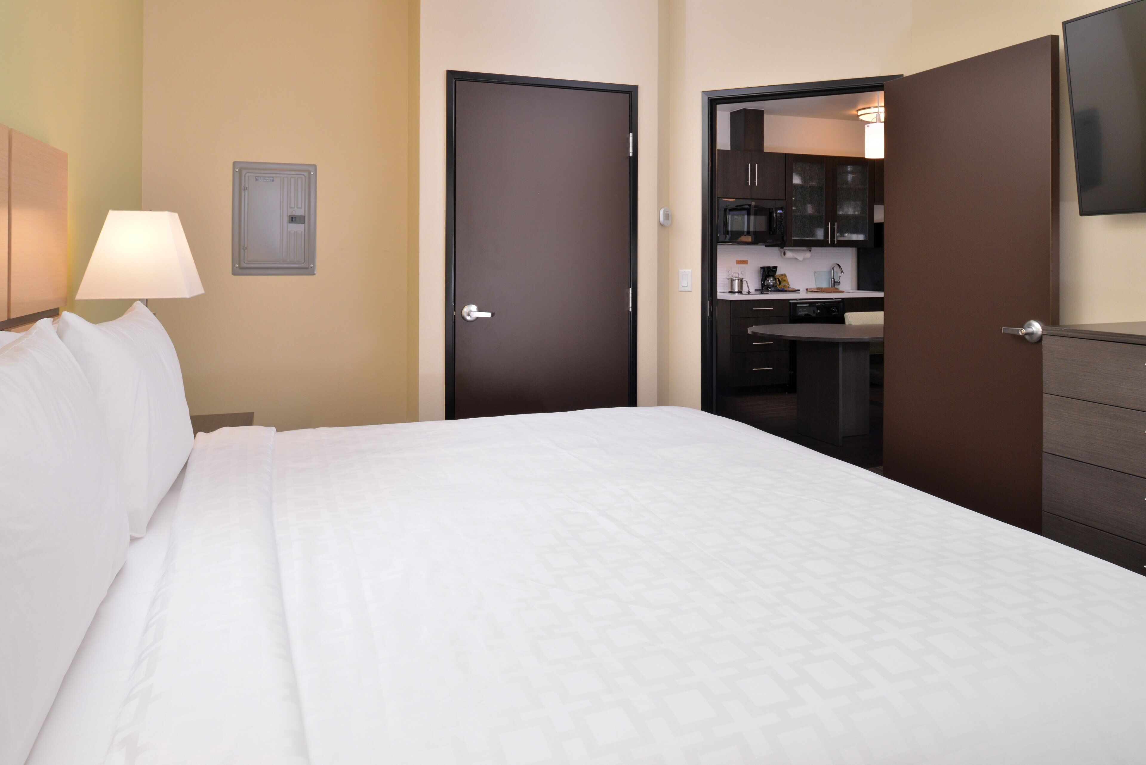 A hotel room with a bed, a nightstand with a lamp, a door leading to a kitchenette, and a wall-mounted TV.