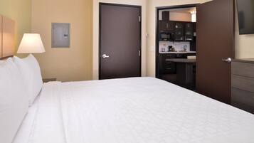A hotel room with a bed, a nightstand with a lamp, a door leading to a kitchenette, and a wall-mounted TV.