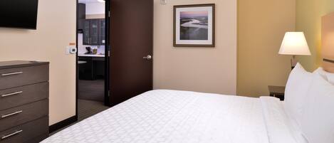 A hotel room with a bed, a dresser, a TV mounted on the wall, a framed picture, and a nightstand with a lamp.