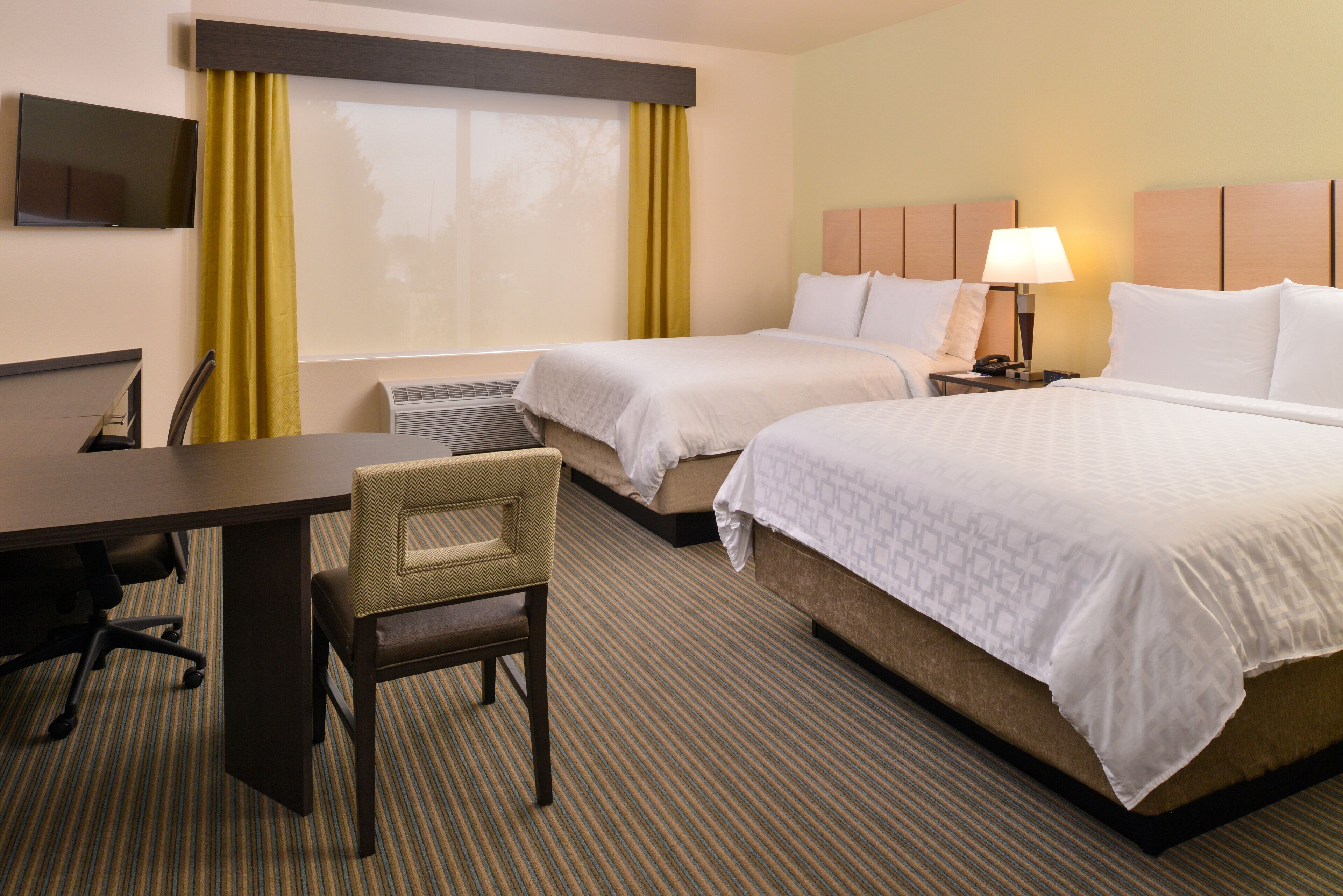 A hotel room with two beds, a desk, a chair, a TV, and a lamp.