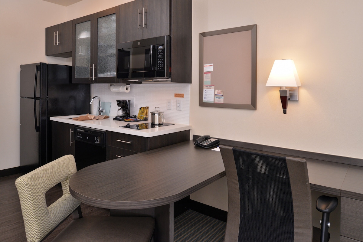A compact kitchen with a refrigerator, microwave, and dishwasher, a dining table with chairs, and a wall-mounted lamp.