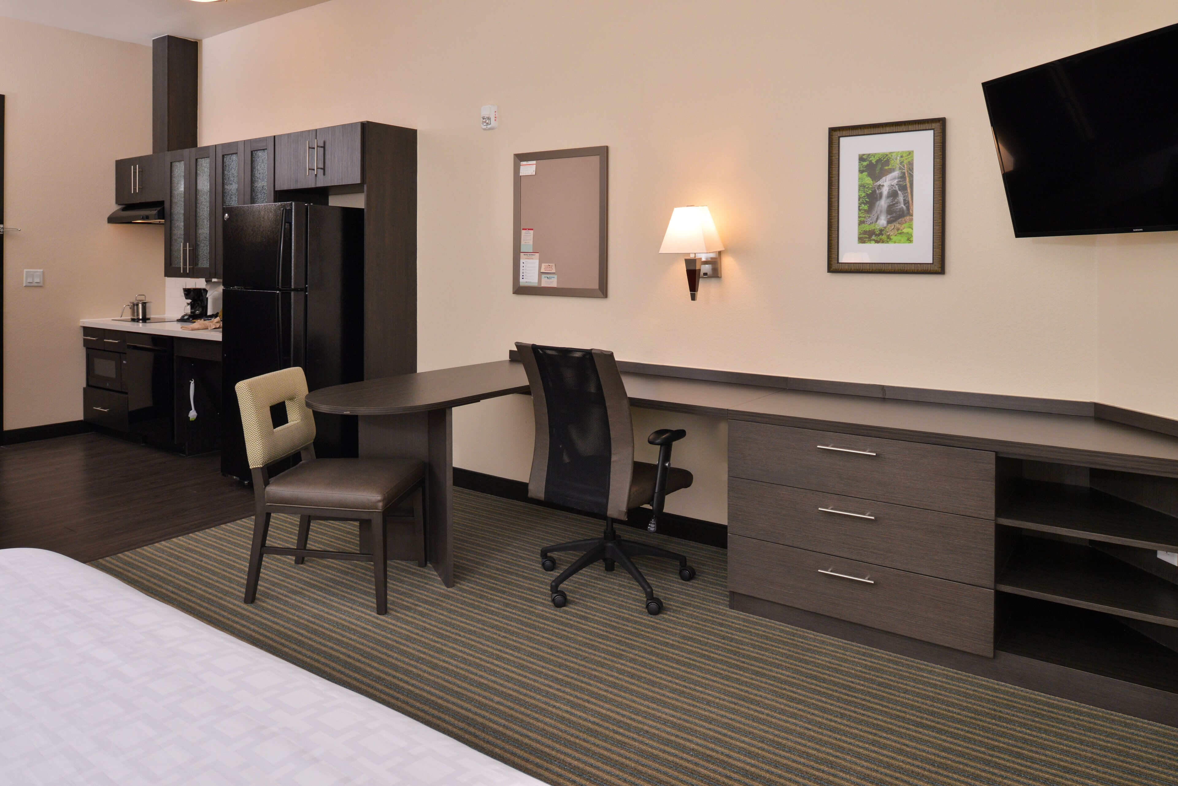 A hotel room with a bed, two chairs, a desk, a refrigerator, a microwave, a sink, and a TV mounted on the wall.