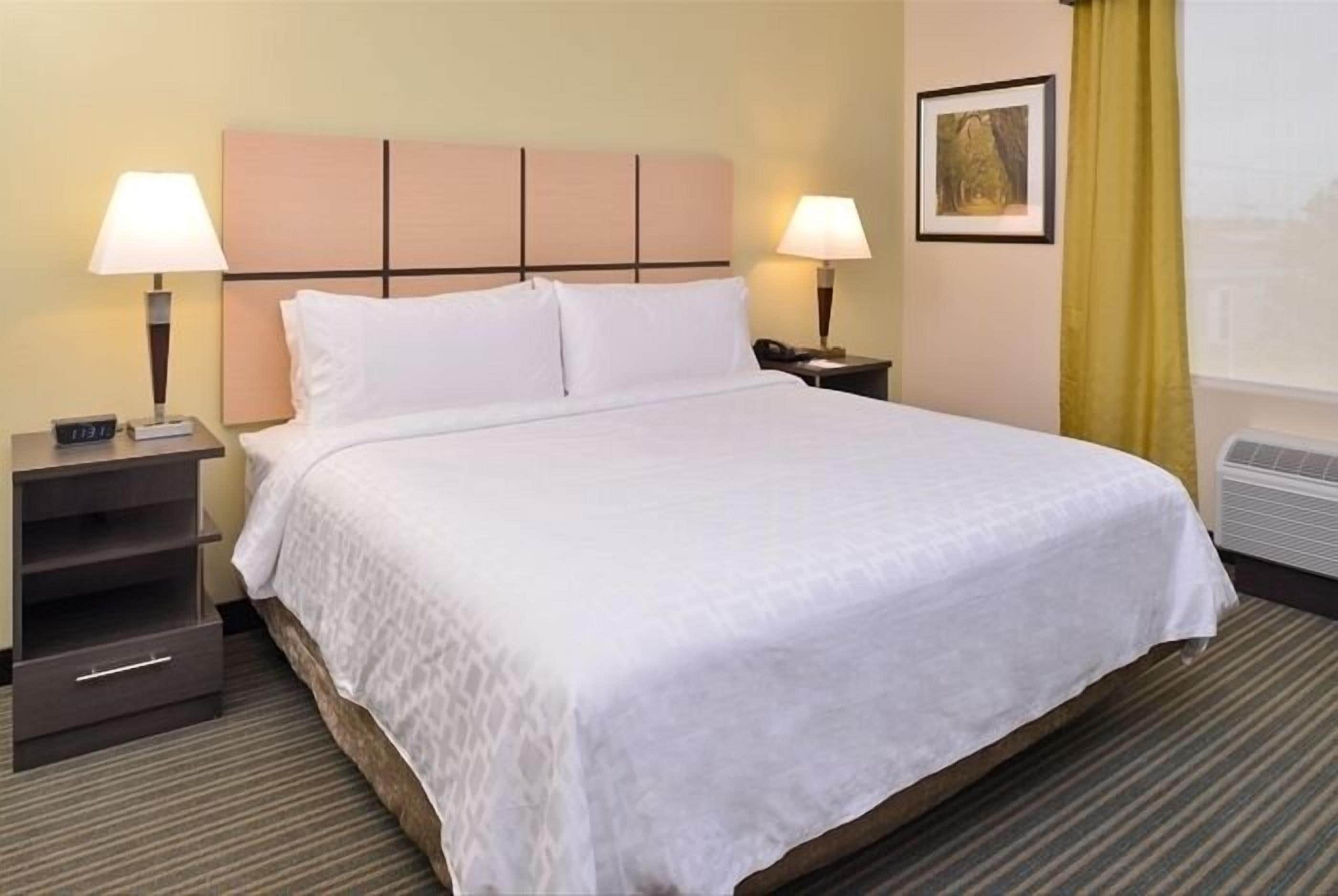 A hotel room with a large bed, two bedside tables with lamps, a small drawer unit, and a framed picture on the wall.
