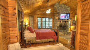 Une chambre située dans un chalet en rondins, avec un grand lit, un plafond en bois, une cheminée en pierre, une télévision et une baignoire.
