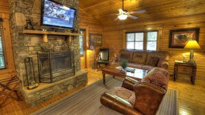 L’intérieur d’un chalet confortable, avec des murs en rondins de bois, une cheminée en pierre, des meubles en cuir et un téléviseur fixé au mur.