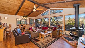 A cozy cabin interior with a fireplace, wooden furniture, and large windows. - Walk to Bear Mtn, National Forest, Golf Course & Zoo! PET FRIENDLY! Outdoor HOT TUB! Amazing Ski Slope Views! (Big Bear Lake)