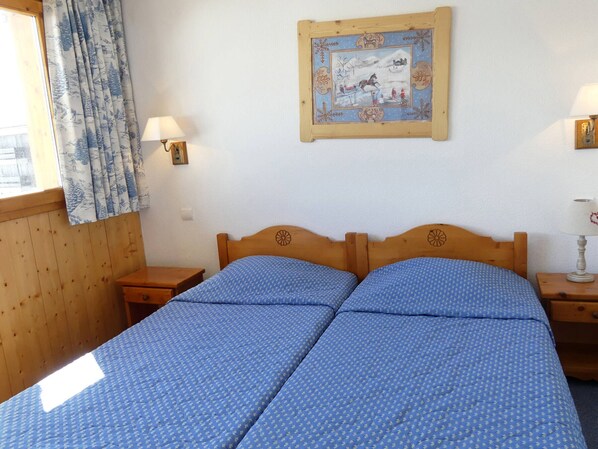 A room with two wooden beds, a window with curtains, and a framed picture on the wall.