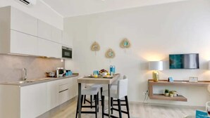 A modern kitchen with white cabinets, a microwave, and a dining area with two chairs. There is a TV mounted on the wall and a shelf with decorative items.