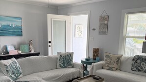 Un salon comprenant un canapé d’angle, une table basse et un ventilateur de plafond. On y trouve une porte avec une fenêtre, un mur orné d’un tableau et une petite table sur laquelle sont disposés des objets décoratifs.