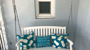 Une balançoire blanche, placée sur un porche, avec deux coussins turquoise ornés de motifs d’ananas et de texte.