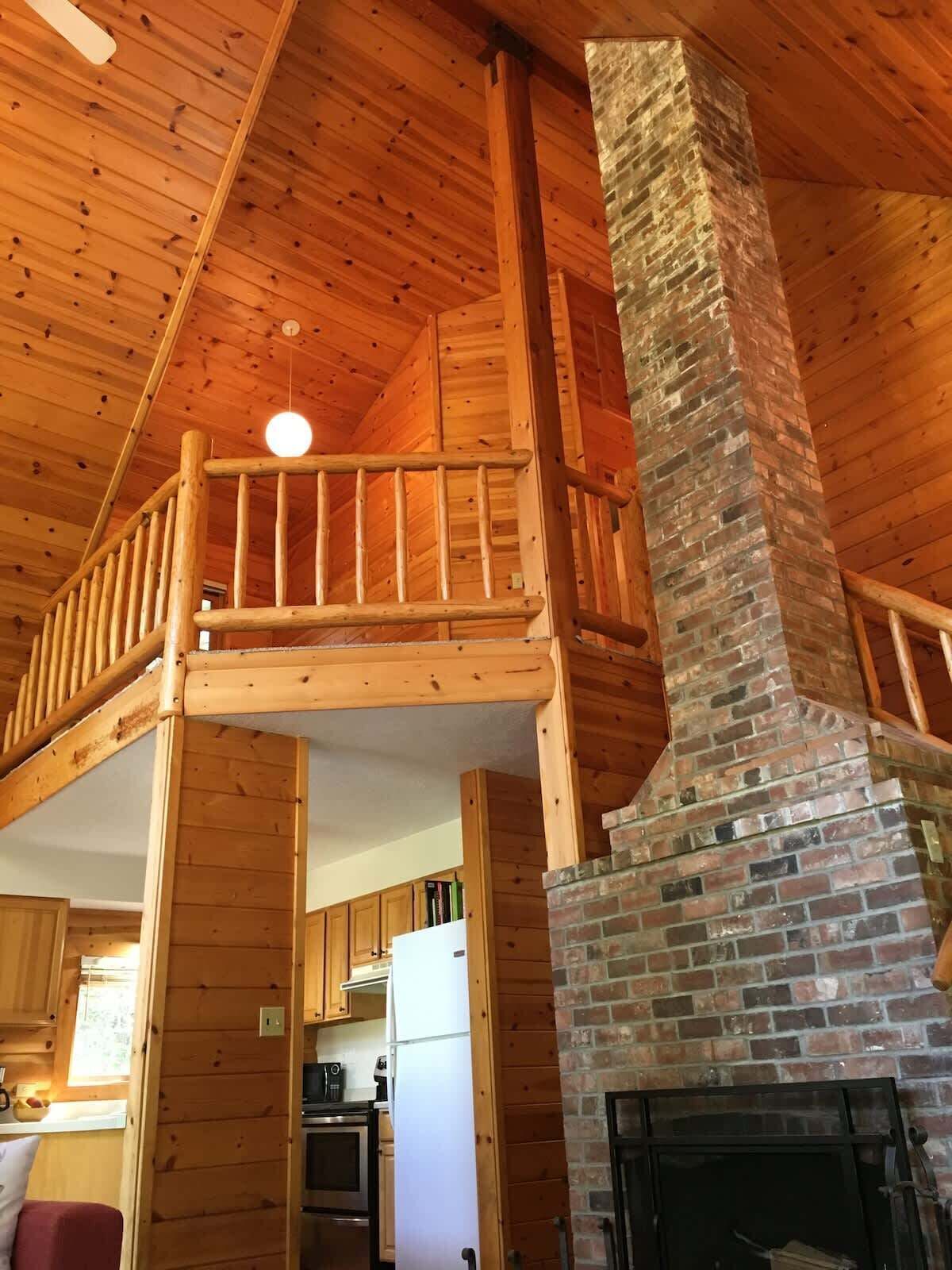 A wooden cabin interior with a fireplace, a balcony, and a kitchen area.