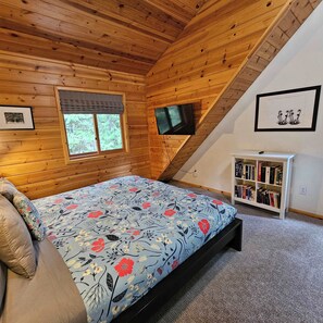 A bedroom with a bed, a window, a TV, a bookshelf, and wooden walls.