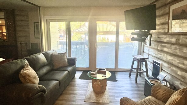 A living room with a leather sofa, a coffee table, a television on the wall, and a view through large windows.