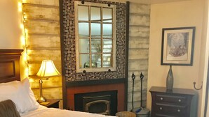 A hotel room with a large bed, a fireplace, a nightstand with a lamp, a framed picture on the wall, and a small table with a vase.