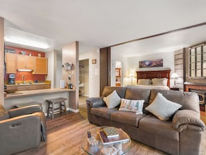 A modern living room with a leather sofa, a glass coffee table, and a kitchen area with wooden cabinets and a countertop.